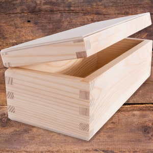 May include: A natural wood box with a hinged lid. The box is made of light wood and has a simple, rectangular shape. The lid is open and the inside of the box is visible.