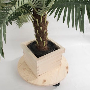 May include: A potted artificial palm tree on a wooden rolling stand. The palm tree has a brown trunk with green leaves. The stand is made of natural wood and has wheels.