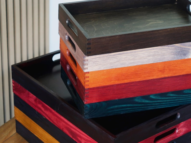 May include: A stack of five wooden trays in various colors, including black, red, orange, gray, and teal. The trays are all rectangular and have a smooth finish.