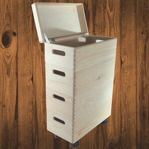 Könnte beinhalten: Eine hohe, rechteckige Holz-Aufbewahrungsbox mit einem Klappdeckel, der geöffnet ist, um das Innere freizugeben. Die Box hat vier horizontale Handschlitze auf der Vorderseite und steht auf kleinen schwarzen Rollen. Das Holz hat eine natürliche, helle Farbe.