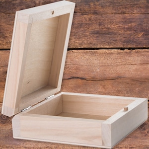 Puede incluir: Una caja de madera vacía con una tapa con bisagras. La caja está hecha de madera de color claro y tiene un acabado natural. La tapa está abierta y el interior de la caja es visible.