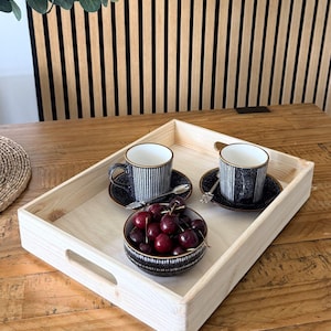May include: A rectangular light-colored wooden serving tray with cutout handles containing two black and white striped ceramic mugs on matching saucers, two small silver spoons, and a small bowl filled with fresh red cherries.