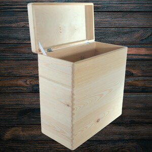 Könnte beinhalten: Eine hellfarbene Holzkiste mit einem Scharnierdeckel, der geöffnet ist, um das Innere freizugeben. Die Kiste ist aus Holzbrettern gefertigt, mit sichtbarer Maserung und natürlicher Oberfläche. Der Deckel ist leicht geöffnet.
