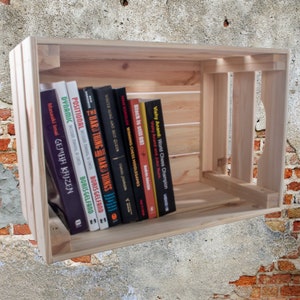 Può includere: Una cassa di legno naturale piena di libri. I libri hanno titoli come "Hard Things" e "Black is still OKI". La cassa è in legno non trattato ed è appoggiata a un muro strutturato.