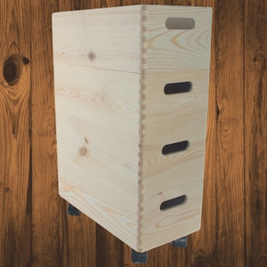 Op de afbeelding: Een hoge, lichtgekleurde houten opbergdoos met vingerlas hoeken en uitgesneden handgrepen. De doos staat op kleine zwarte wielen en heeft een natuurlijke houtnerf afwerking. De doos is ontworpen voor opslag.