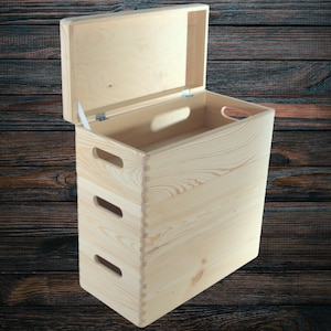 May include: A light-coloured wooden storage box with a hinged lid, open to reveal the interior. The box has two handles on the sides and three cut-out handles on the front. The wood grain is visible, and the box appears to be new.