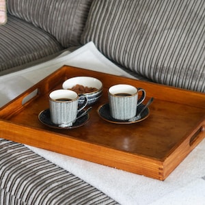 May include: A wooden tray with two black and white striped coffee mugs and a small bowl of nuts. The tray is sitting on a white blanket on a gray couch.