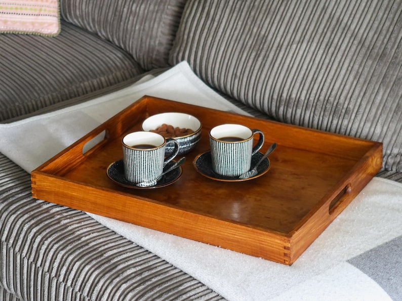 May include: A wooden tray with two black and white striped coffee mugs and a small bowl of nuts. The tray is sitting on a white blanket on a gray couch.