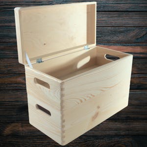 May include: A light-coloured wooden storage box with a hinged lid, open to reveal the interior. The box has cut-out handles on the sides and is constructed with dovetail joints. The background is a dark wood grain.