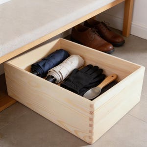 Può includere: Una scatola rettangolare in legno chiaro con angoli a incastro. All'interno della scatola ci sono due ombrelli piegati, uno blu navy e uno beige, un paio di guanti neri e un piccolo cucchiaio in metallo con un manico in legno chiaro.