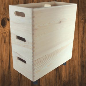 Op de afbeelding: Een hoge, rechthoekige houten opbergbak met drie handgreepuitsparingen aan de voorkant. De bak is gemaakt van lichtgekleurd hout, dat de natuurlijke nerf laat zien. Hij staat op kleine zwarte wielen, wat mobiliteit suggereert. Het ontwerp is eenvoudig en functioneel.