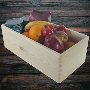 Op de afbeelding: Een houten kist gevuld met verse producten en conserven. De kist bevat een banaan, sinaasappels, appels, tomaten en conserven. De kist is gemaakt van lichtgekleurd hout en staat op een donkere houten ondergrond.
