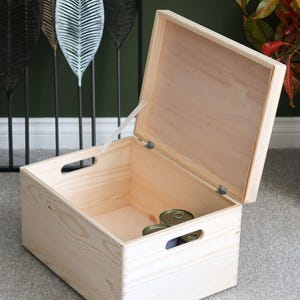 May include: A rectangular light-coloured wooden storage crate with a hinged lid that is propped open. The box features finger-jointed corners and two oval-shaped cut-out handles on the sides. Inside the crate, two small metal cans are visible.