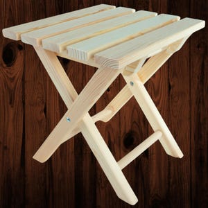 Könnte beinhalten: Ein kleiner, zusammenklappbarer Holzschemel mit natürlicher Oberfläche. Der Hocker hat eine Sitzfläche aus Latten und ein X-Rahmen-Design für Stabilität. Der Hocker ist ca. 30 cm lang, 25 cm breit und 25 cm hoch, wenn er geöffnet ist.