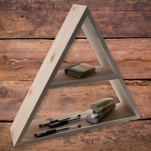 May include: A light-colored, triangular wooden shelf with two levels. The top shelf holds a small notepad with colorful sticky notes. The bottom shelf displays two black pens and a green stapler. The shelf is set against a wooden background.