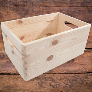 Puede incluir: Una caja de almacenamiento rectangular de madera sin terminar con construcción de ensamblaje de dedos. La caja tiene asas recortadas en los lados y está hecha de madera de color claro con vetas y nudos visibles. La caja está sobre una superficie de madera.