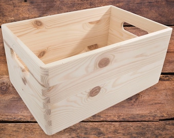 Peut inclure: Une caisse de rangement rectangulaire en bois non fini avec une construction à joints à doigts. La caisse a des poignées découpées sur les côtés et est faite de bois de couleur claire avec des veines et des nœuds visibles. La caisse repose sur une surface en bois.