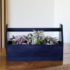 May include: A dark blue wooden tool box planter with a handle. The planter is filled with green and purple plants.