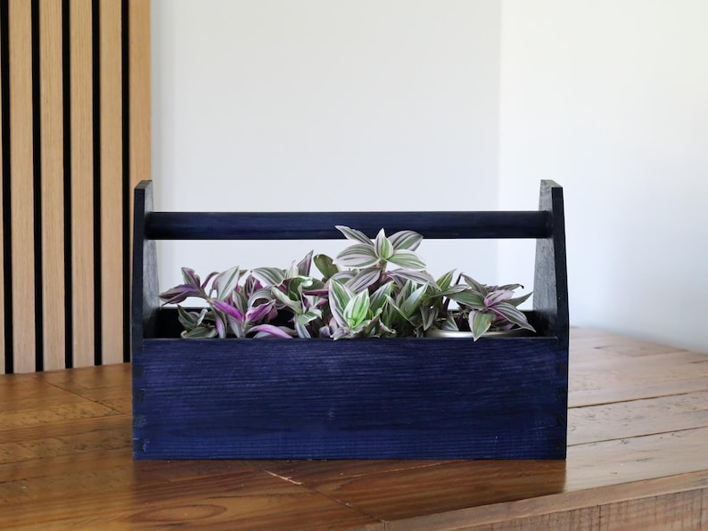 May include: A dark blue wooden tool box planter with a handle. The planter is filled with green and purple plants.