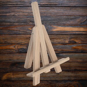 May include: A small wooden easel made of light brown wood. The easel is designed to hold a small painting or photograph.