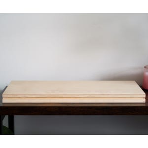 May include: Two light-colored wooden boards stacked on top of each other. The boards are rectangular and appear to be made of plywood. The boards are sitting on a dark brown table, with a white wall in the background.