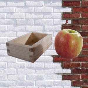 May include: A wooden rectangular box with a natural wood finish. The box is empty and has a smooth surface. The box is sitting on a white brick wall. A red apple is sitting on a red brick wall to the right of the box.