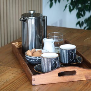 May include: A wooden tray with a handle holds a silver French press, two black and white striped mugs, a small white creamer, a bowl of cookies, and a glass of water.