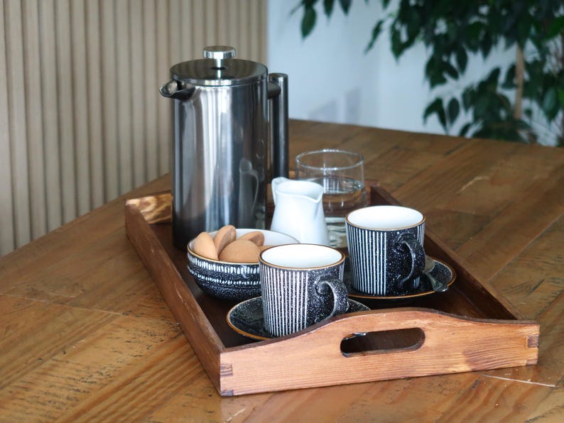 May include: A wooden tray with a handle holds a silver French press, two black and white striped mugs, a small white creamer, a bowl of cookies, and a glass of water.