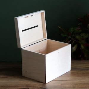 May include: A light-colored wooden box with a hinged lid, open to reveal the interior. The lid has a slot cut into it. The box is sitting on a wooden surface, with a dark green background.