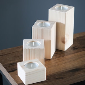 May include: Four wooden candle holders of varying sizes, each with a small white tea light candle inside. The holders are arranged in a row on a wooden table, with the largest holder at the back and the smallest holder at the front.