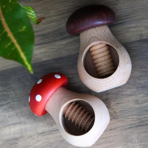 Op de afbeelding: Twee houten paddenstoelvormige notenkrakers. Eén heeft een rode kap met witte stippen, de andere een donkerbruine kap. Beide hebben een licht houten lichaam met een zichtbaar schroefmechanisme voor het kraken van noten. Op een houten oppervlak.