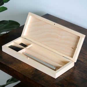 May include: An open, light-colored wooden pen box with a silver pen inside. The box has a small compartment for other items. The box is on a dark brown wooden surface.