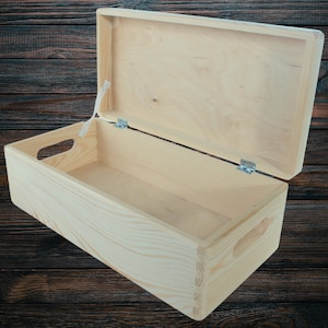 Pode incluir: Uma caixa de armazenamento de madeira natural com tampa articulada e duas alças nas laterais. A caixa está aberta e vazia.