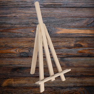 May include: A wooden easel with a triangular base and three legs. The easel is made of light-colored wood and is designed to hold a small canvas or painting.