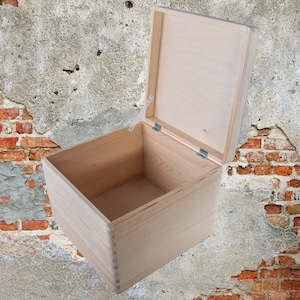 May include: An empty wooden box with a hinged lid. The box is made of light-coloured wood and has a natural finish. The lid is open and the box is sitting on a textured surface.