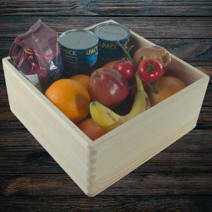 May include: A wooden crate filled with fresh produce and canned goods. The crate contains oranges, apples, tomatoes, a banana, and two cans. A bag of coffee is also visible. The crate is made of light-colored wood.