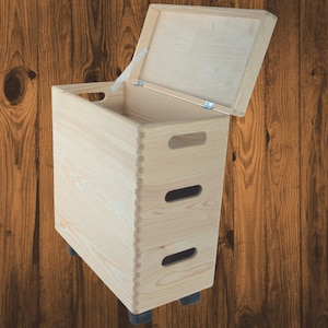 Könnte beinhalten: Eine hellfarbene Holz-Aufbewahrungsbox mit einem Scharnierdeckel, der geöffnet ist, um das Innere freizulegen. Die Box hat zwei Griffaussparungen und rollt auf kleinen schwarzen Rädern. Die Holzmaserung ist sichtbar.
