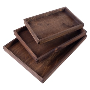 Può includere: Tre vassoi di legno di diverse dimensioni impilati uno sopra l'altro. I vassoi sono in legno marrone scuro e hanno un aspetto rustico.