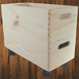 Puede incluir: Una caja de almacenamiento de madera de color claro con esquinas ensambladas con dedos y asas recortadas. La caja está montada sobre cuatro pequeñas ruedas negras para facilitar el movimiento. La veta de la madera es visible.