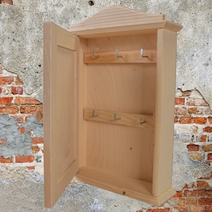 Könnte beinhalten: Ein Schlüsselhalter aus Holz mit einer Flügeltür und mehreren Haken. Der Schlüsselhalter ist aus hellem Holz gefertigt und hat ein schlichtes Design.