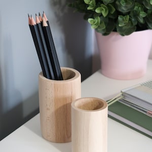 May include: Two wooden pencil holders, one larger than the other, with a set of black pencils in the larger holder. The holders are on a white surface.