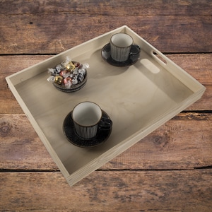 Op de afbeelding: Een licht houten dienblad met twee zwart-wit gestreepte koffiekopjes en schotels. Een kleine kom met verpakte snoepjes staat op het dienblad. De lade heeft uitgesneden handgrepen en staat op een houten oppervlak.