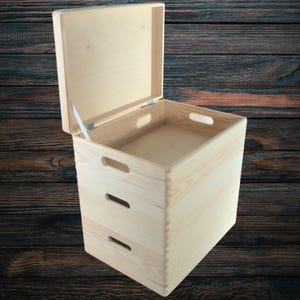 May include: A light-coloured wooden storage box with a hinged lid, open to reveal the interior. The box has two cut-out handles on the sides and a third on the front. The box is made of unfinished wood.
