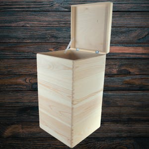 May include: A tall, unfinished wooden box with a hinged lid, open to reveal the interior. The box is constructed from light-coloured wood, showcasing the natural grain. The lid is slightly ajar, revealing the interior space. The box is set against a dark wood background.