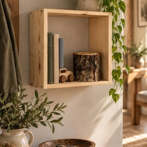 May include: A square, light-coloured wooden wall shelf mounted on a wall. Inside the shelf are three vertical books, a small wooden toy car, and a cylindrical candle with a textured, bark-covered exterior. The wood grain is visible throughout.
