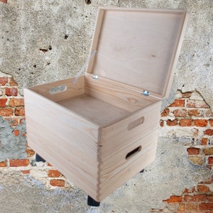 Può includere: Una scatola di legno naturale con un coperchio incernierato e due maniglie sul davanti. La scatola è aperta e si trova su un muro di mattoni.