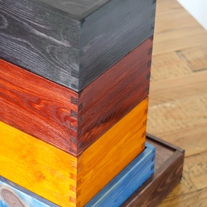 May include: A stack of four wooden boxes with different colored stains. The top box is black, the second is red, the third is yellow, and the bottom box is blue. The boxes are stacked on a wooden surface.