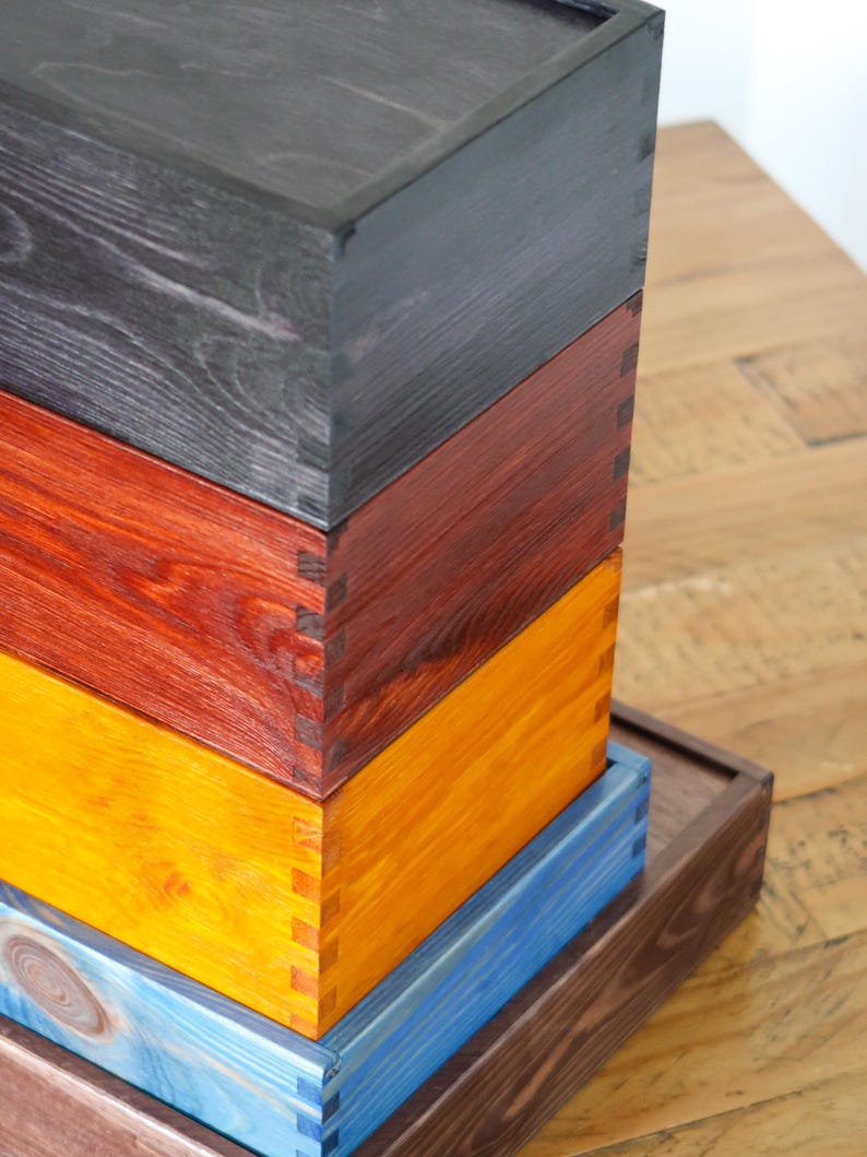 May include: A stack of four wooden boxes with different colored stains. The top box is black, the second is red, the third is yellow, and the bottom box is blue. The boxes are stacked on a wooden surface.