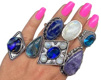 Puede incluir: Una colección de anillos de piedras preciosas en varias formas y colores, incluyendo piedras azules, moradas y transparentes. Los anillos están engarzados en metal plateado con diseños intrincados. Los anillos se muestran en una mano con esmalte de uñas rosa brillante.