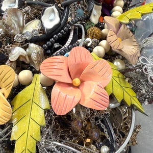 May include: A jumble of jewellery items, including necklaces, bracelets, and earrings. The collection features silver chains, beaded strands, and pendants. Wooden flower and leaf accents in peach and yellow add colour and natural elements to the mix.